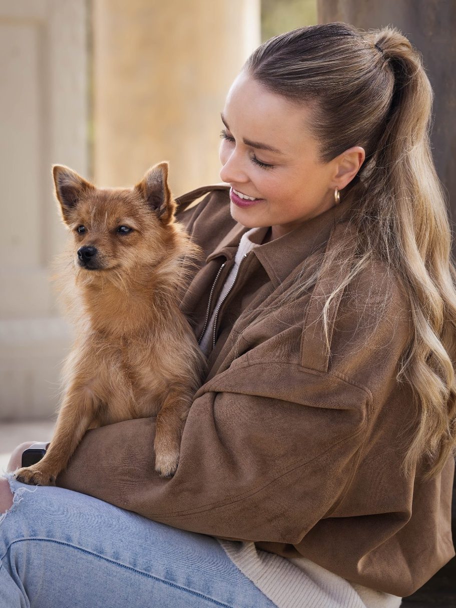 Junge Frau mit langen Haaren hält einen kleinen, braunen Hund auf dem Arm.