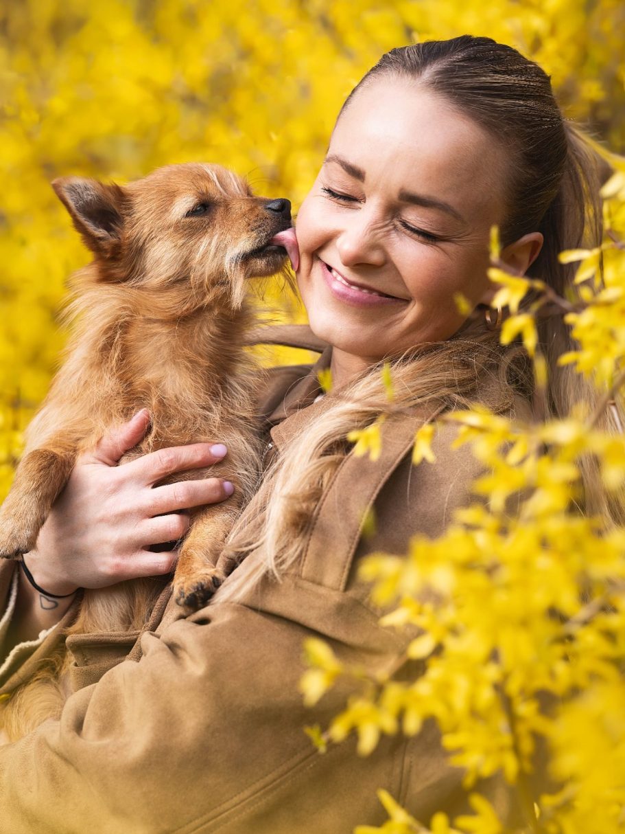 Frau umarmt einen kleinen Hund, umgeben von gelben Blumen. Beide lächeln glücklich.